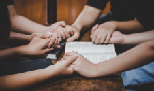 People holding hands praying over a Bible