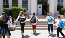 Five children wearing backpacks running up the stairs into a school