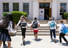 Five children wearing backpacks running up the stairs into a school